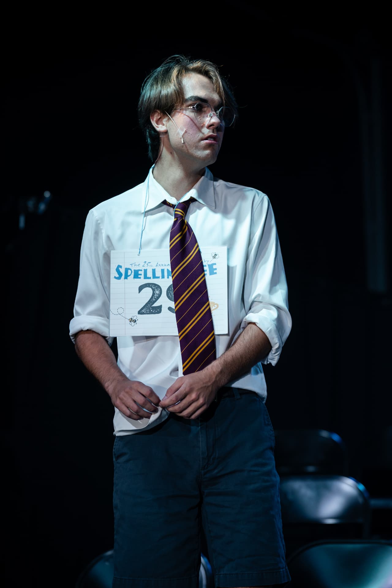 Richard Jordan standing with his number 29 card in a moody spotlight during Putnam County Spelling Bee