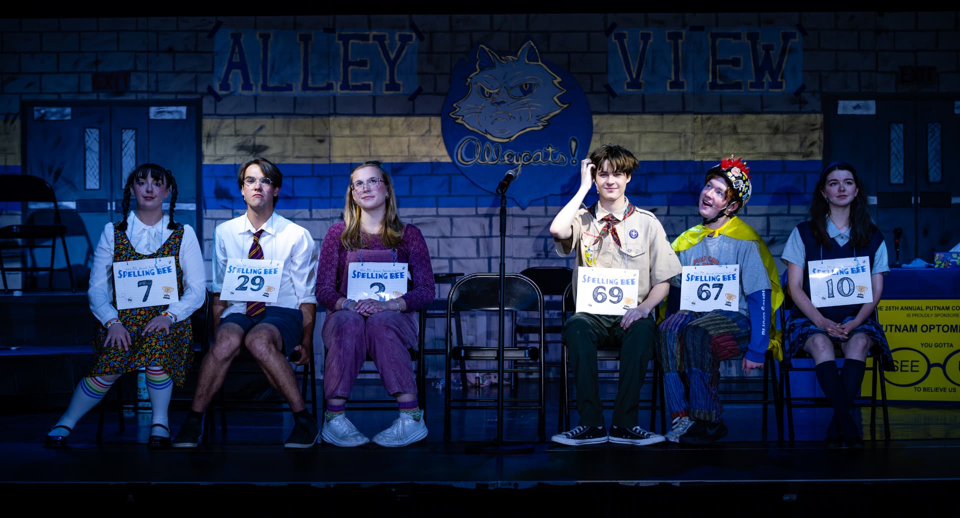 Richard Jordan center stage with the seated spellers on the Alleycats gym set of Putnam County Spelling Bee