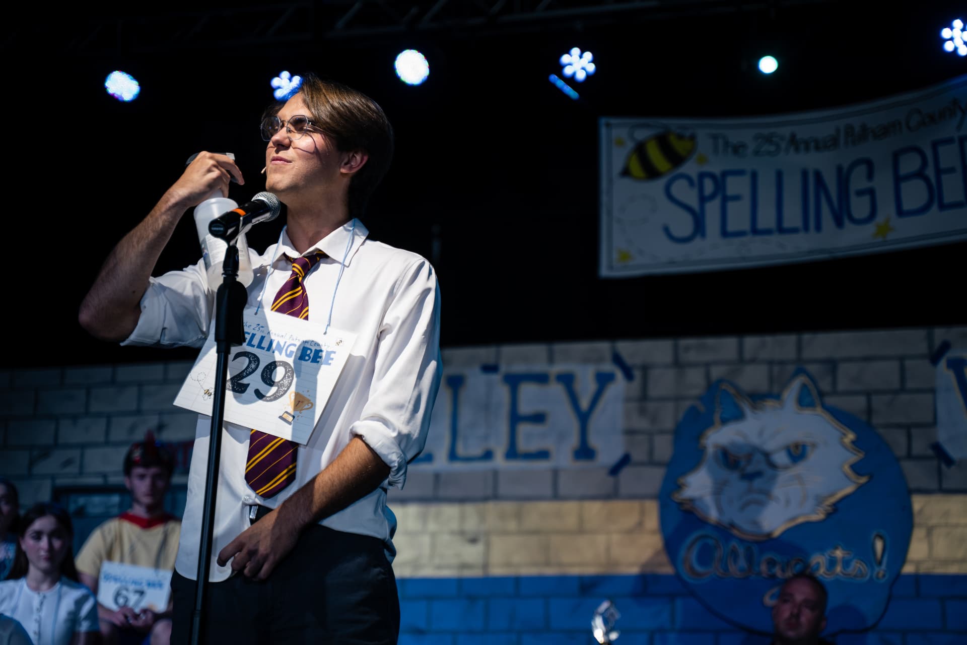 Richard Jordan at the microphone raising his spray bottle beneath the Spelling Bee banner