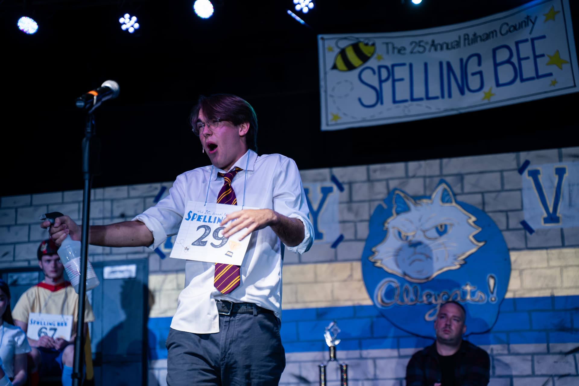 Richard Jordan mid-song at the microphone with his spray bottle outstretched beneath the Spelling Bee banner