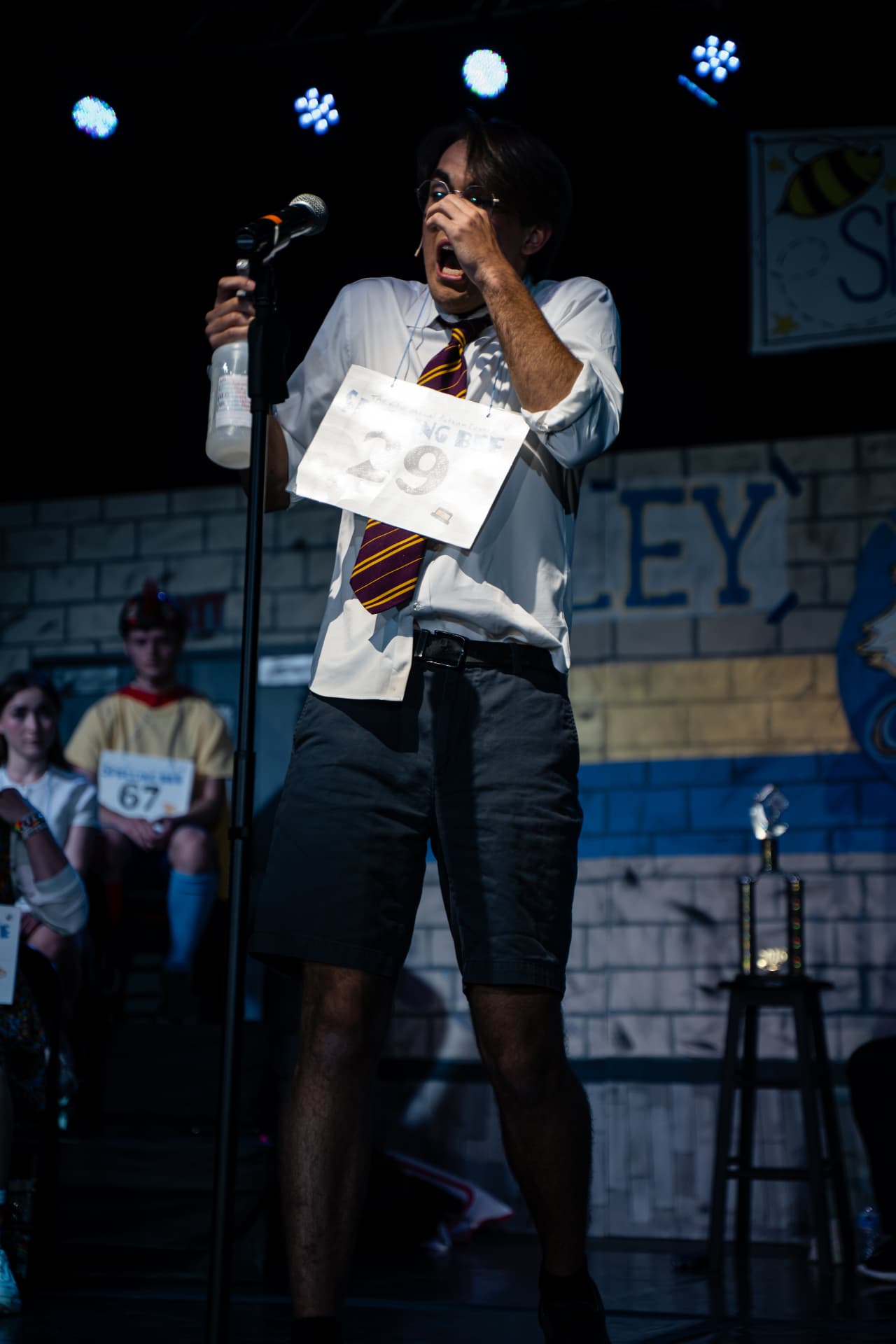 Richard Jordan singing at the microphone holding a spray bottle in Putnam County Spelling Bee