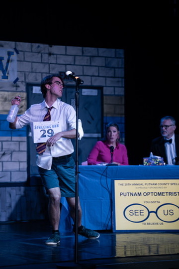 Richard Jordan at the microphone gesturing with a spray bottle as the judges watch
