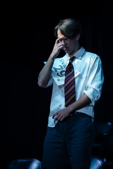 Richard Jordan adjusting his glasses in a dark spotlight during Putnam County Spelling Bee