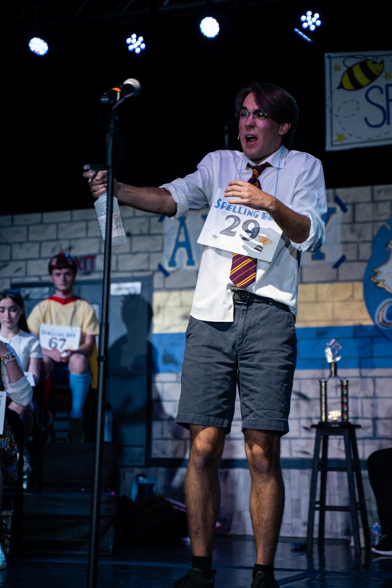 Richard Jordan with arms outstretched holding his spray bottle on the Spelling Bee gymnasium set
